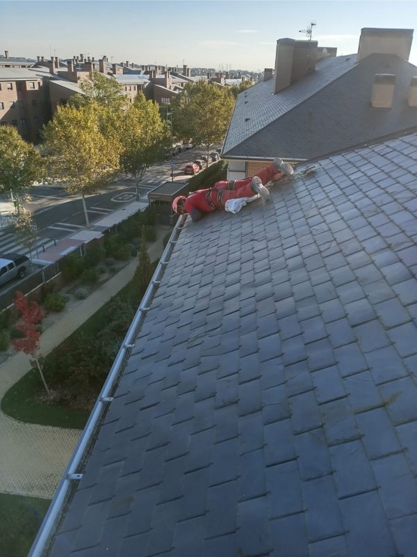 Operario en equipo de seguridad rojo realizando labores de inspección y mantenimiento en la cubierta de pizarra de un edificio, asegurado con arnés de seguridad, en un entorno urbano residencial durante un día soleado.