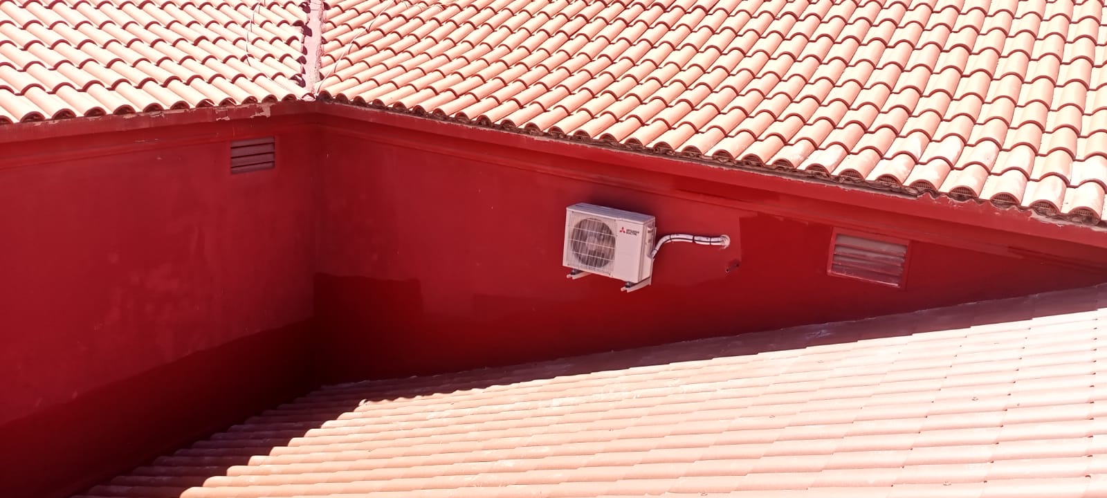 Vista de un tejado de tejas rojas con una unidad de aire acondicionado en una pared roja, ilustrando las labores de modernización y mantenimiento en el contexto de un proyecto de rehabilitación de edificios para mejorar la funcionalidad y eficiencia energética sin alterar su estética tradicional.