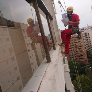 Imagen vertical de un trabajador colgado con arneses de seguridad mientras realiza una inspección técnica en la fachada de un edificio. La reflexión del entorno urbano en los ventanales del edificio añade profundidad a la escena, enfatizando la altura y la precisión requerida en el mantenimiento y rehabilitación de edificaciones urbanas.