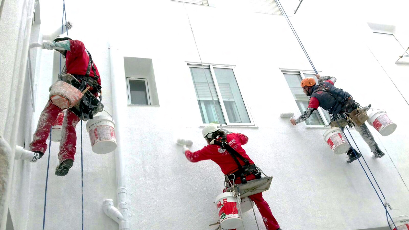 Operarios vestidos con equipo de seguridad completo y arneses, realizando trabajos de pintura en la fachada blanca de un edificio residencial, empleando técnicas de escalada y acceso por cuerda para la rehabilitación urbana.