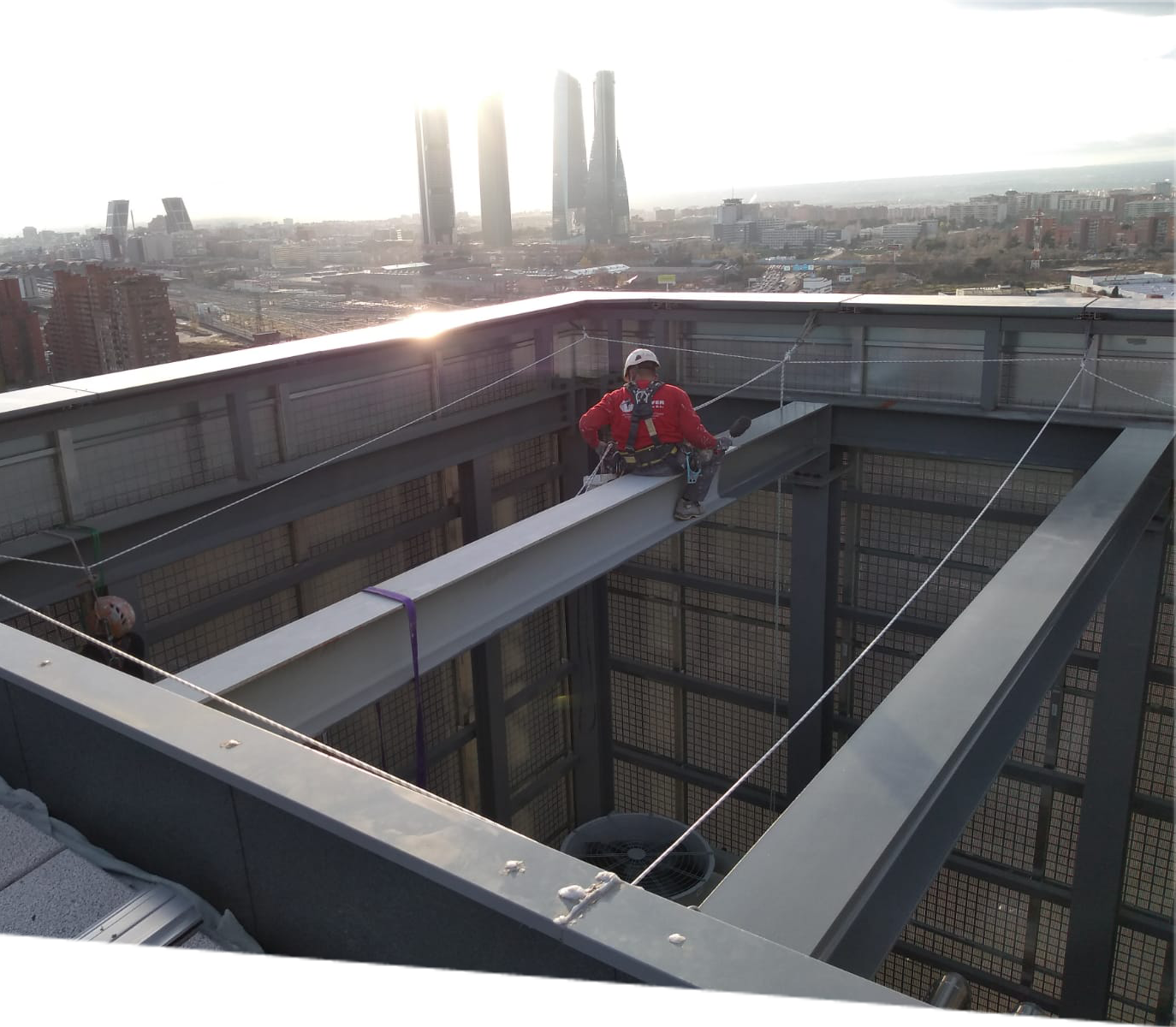 Operario de construcción en equipo de seguridad rojo asegurado con arnés mientras realiza trabajos de mantenimiento en la estructura metálica de un edificio alto, con el horizonte de Madrid y las Cuatro Torres en el fondo en un día soleado.
