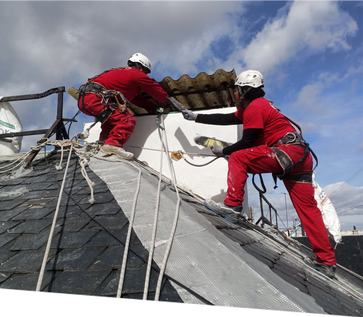 Dos trabajadores con equipos de seguridad realizan trabajos de rehabilitación en el tejado de un edificio histórico, restaurando la estructura y aplicando pintura en un día nublado, enfatizando la conservación del patrimonio arquitectónico.