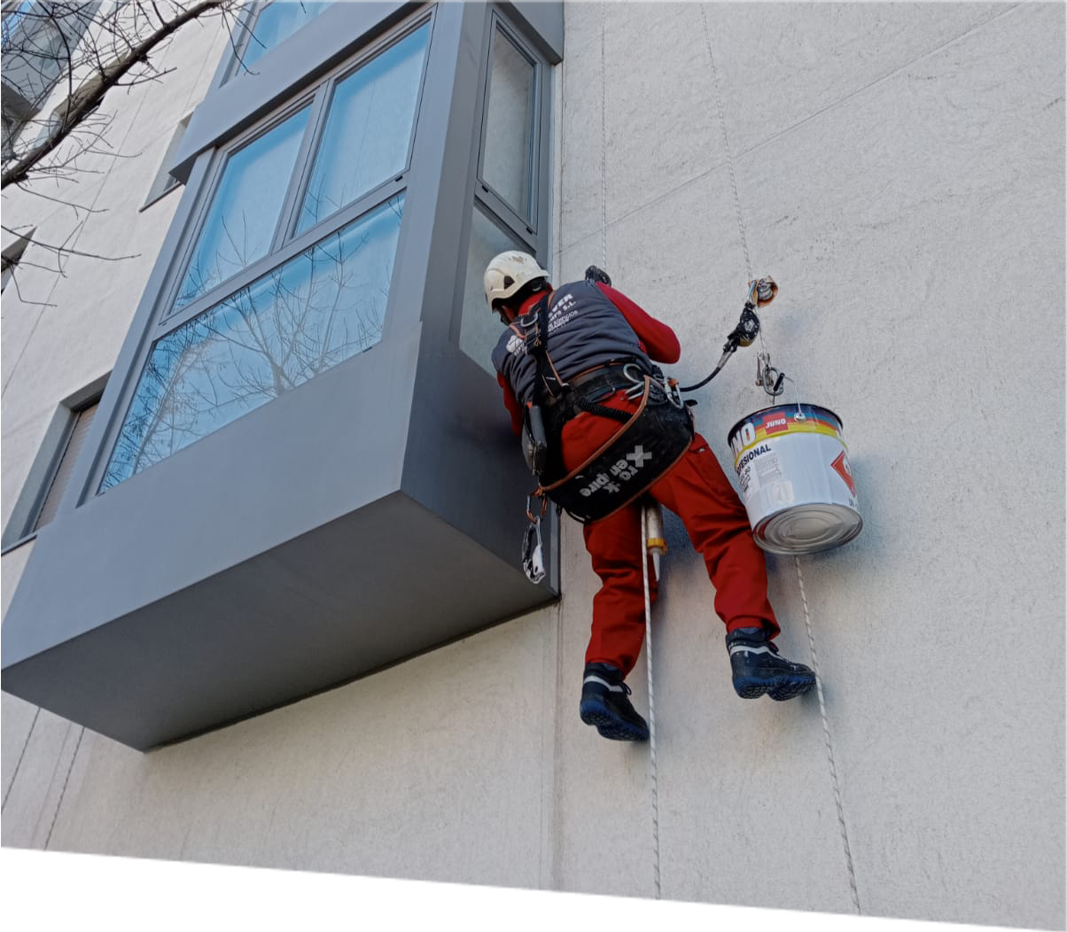 Trabajador con arnés de seguridad realiza trabajos de pintura en la fachada exterior de un moderno edificio urbano, demostrando técnicas avanzadas de rehabilitación arquitectónica en altura en un entorno urbano.